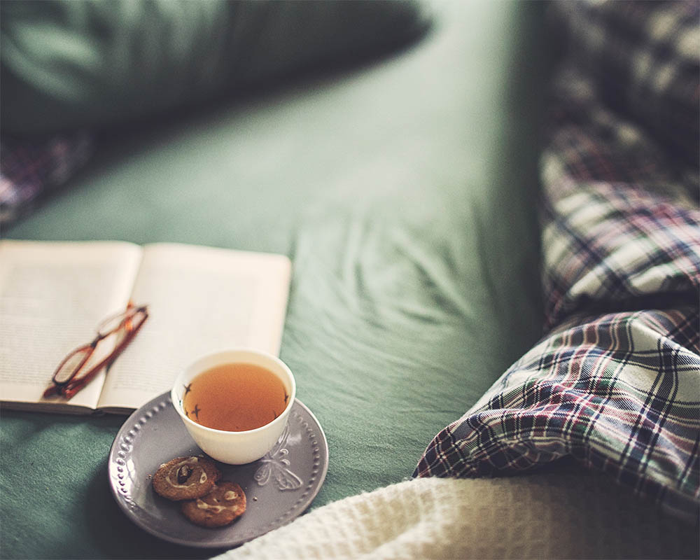 Cosy image of a mug of tea, cookies, a book, and reading glasses in bed.