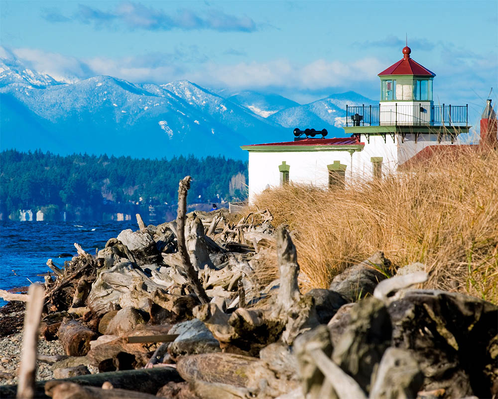 Image of a lighthouse on a rocky beach with snow capped  mountains in the background.