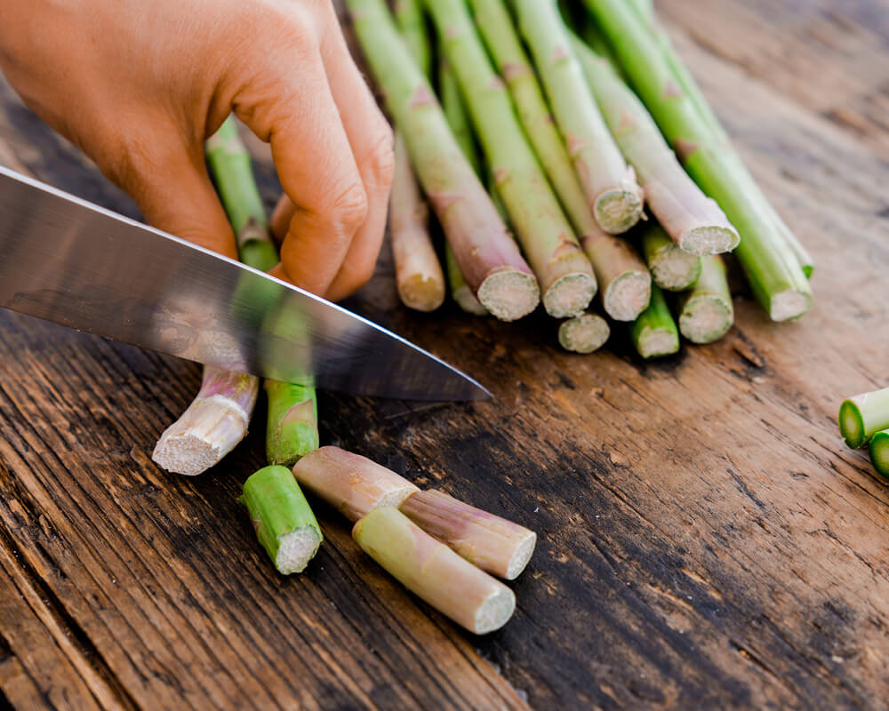 a left hand holds asparagus on a wood surface while a knife trims off the ends